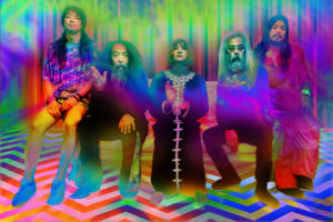 Acid Mothers Temple band photograph. The band is seated on a couch and there is a green purple blue filter over the photograph.